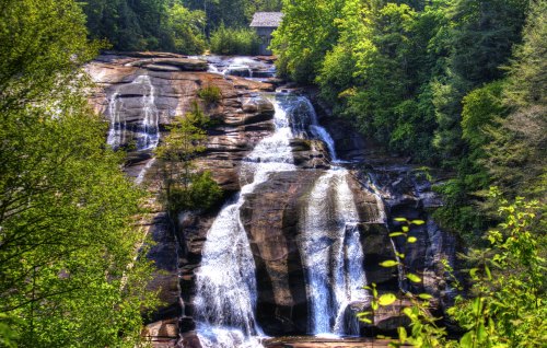 A scenic waterfall cascading over rocky cliffs, surrounded by lush green trees and a distant cabin.