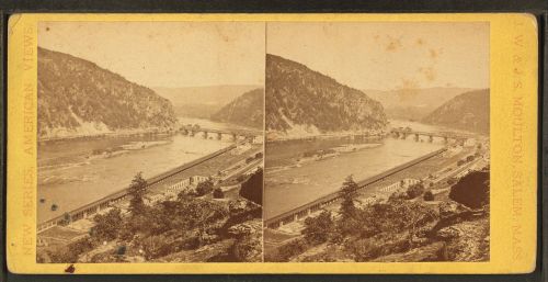 A vintage stereoscopic view of a river valley with a bridge and mountains in the background.