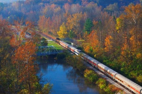 A train travels along a river surrounded by vibrant autumn foliage and trees in a scenic landscape.