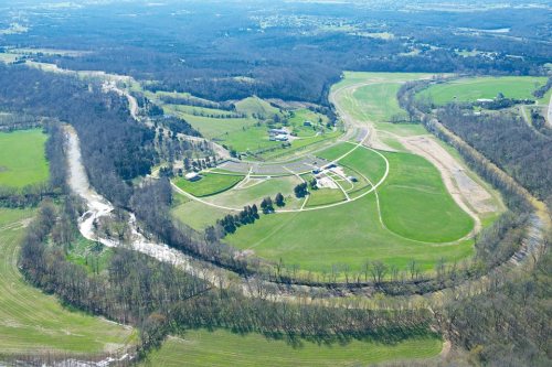 Aerial view of a green landscape with fields, a winding river, and a cluster of buildings surrounded by trees.