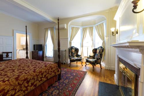 Cozy bedroom with a four-poster bed, two armchairs, a fireplace, and large windows with drapes. Warm, inviting atmosphere.