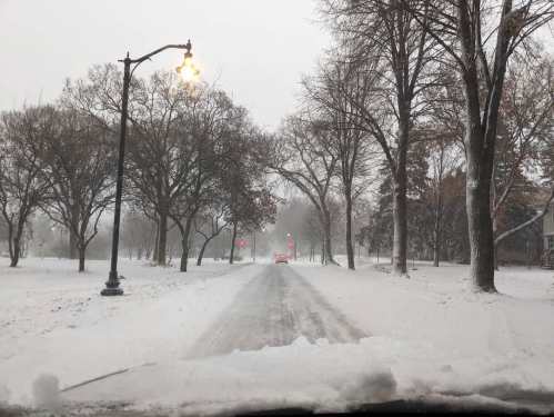 A snowy street lined with bare trees, traffic lights glowing, and a car approaching in the distance.