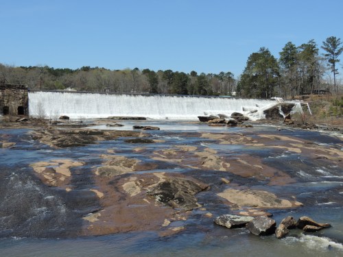 A wide waterfall cascades over a dam, surrounded by rocky terrain and trees under a clear blue sky.