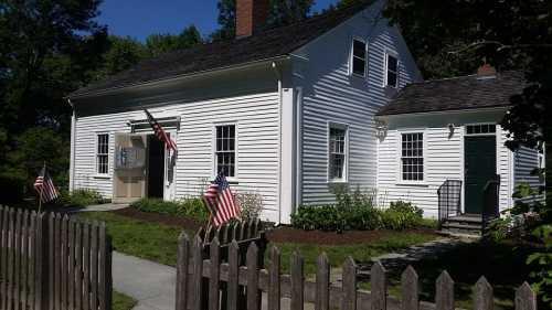 A white, historic house with American flags in front, surrounded by greenery and a picket fence on a sunny day.