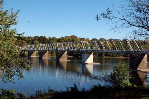 A metal bridge spans a calm river, surrounded by trees with autumn foliage under a clear blue sky.