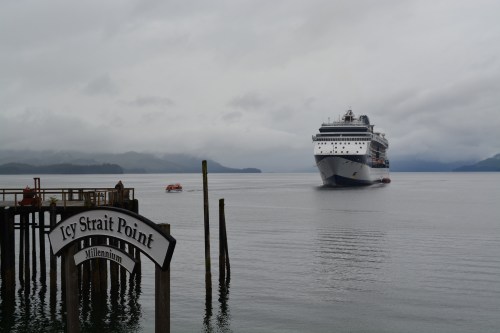 A cruise ship approaches Icy Strait Point under a cloudy sky, with a wooden sign in the foreground.