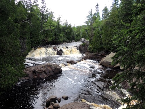 A serene river flows over rocks, surrounded by lush green trees and a cloudy sky.