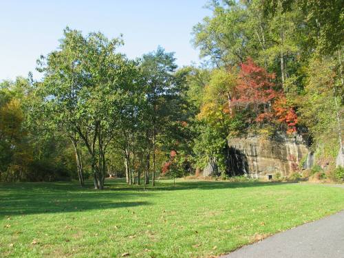 A serene park scene featuring green grass, trees, and a rocky cliff with autumn foliage in the background.