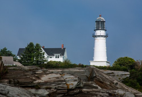 A white lighthouse stands tall beside a house, surrounded by rocky terrain and greenery under a cloudy sky.