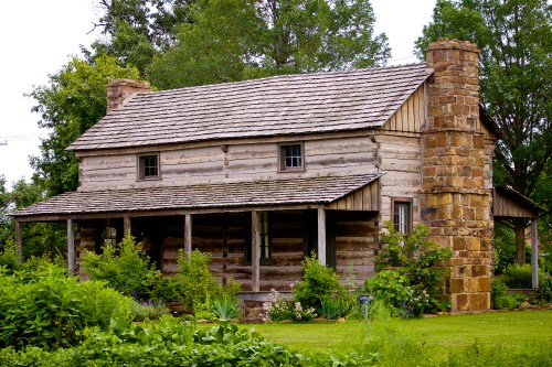 A rustic log cabin with a stone chimney, surrounded by greenery and trees.