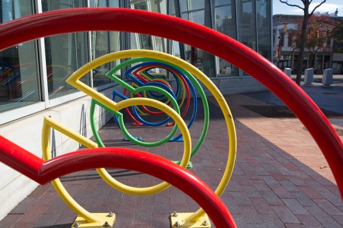 Colorful, curved bike racks in red, yellow, and green arranged in a playful pattern outside a modern building.