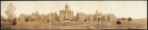 Historic panoramic view of a large, ornate building surrounded by manicured lawns and trees.