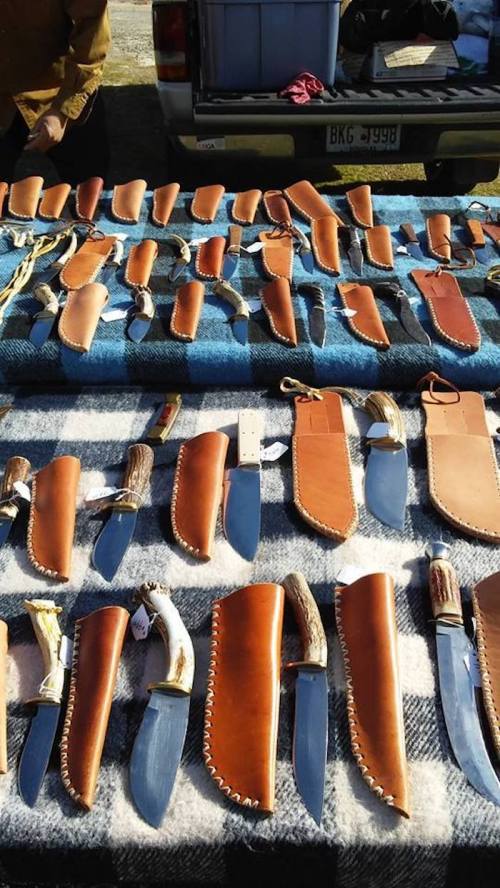A display of various knives with leather sheaths arranged on a table, set against a backdrop of a vehicle.