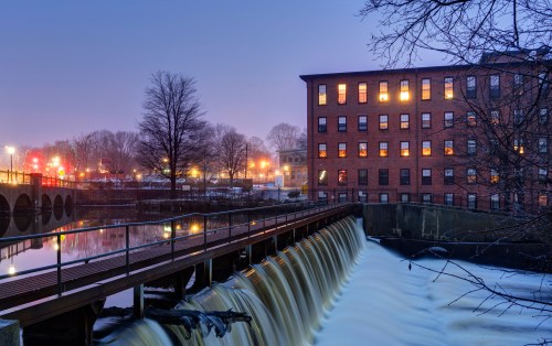 A serene evening scene of a waterfall beside a brick building, with city lights reflecting on the water.