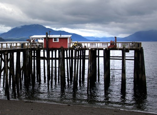 A wooden pier extends over calm water, with a small red building at the end and mountains in the background under cloudy skies.