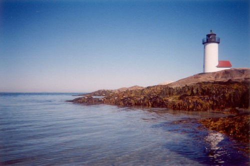 A lighthouse stands on rocky shore, overlooking calm waters under a clear blue sky.