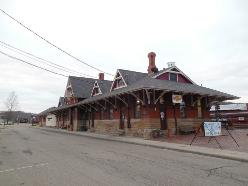 Historic train station with a stone and wood exterior, featuring a sloped roof and decorative eaves, set along a quiet street.