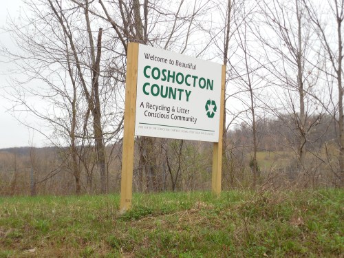 Sign welcoming visitors to Coshocton County, highlighting its recycling and litter-conscious community. Trees in the background.