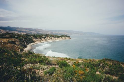 A scenic coastal view with rolling hills, a sandy beach, and gentle waves under a cloudy sky.