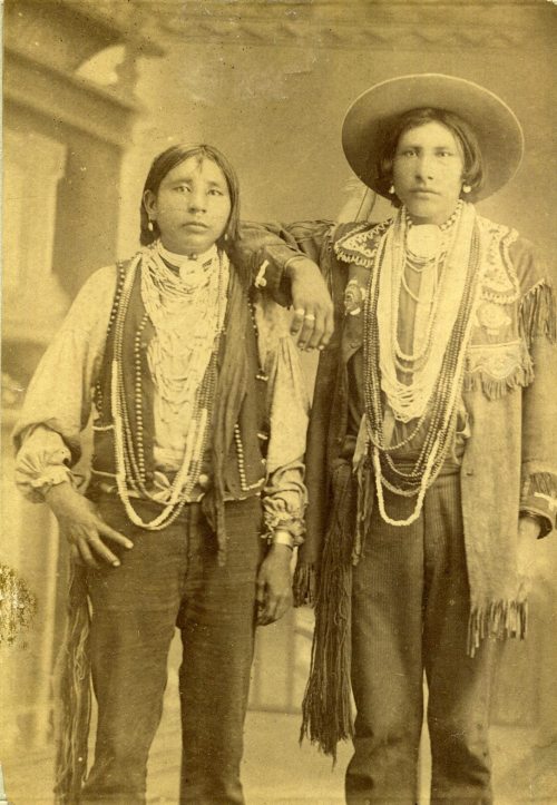 Two Indigenous individuals pose together, adorned in traditional clothing and jewelry, with a vintage sepia tone.