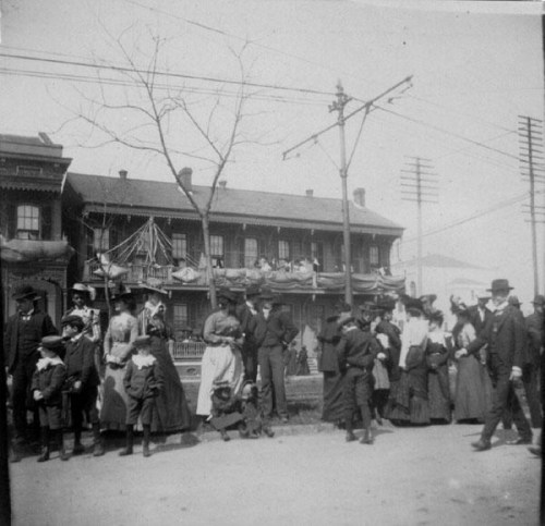 A historic black-and-white photo of a crowd outside a building, with people in period clothing and utility poles in the background.