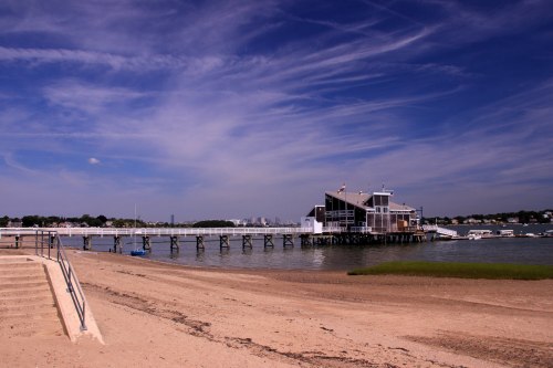 A scenic view of a pier and waterfront building under a blue sky with wispy clouds, alongside a sandy beach.