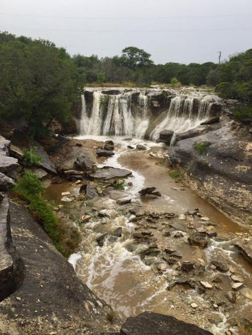 A scenic view of a waterfall cascading over rocky terrain, surrounded by greenery and a flowing river below.