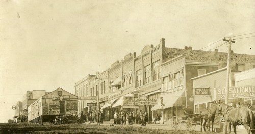 Historic street scene featuring early 20th-century buildings, horse-drawn carriages, and people gathered outside shops.
