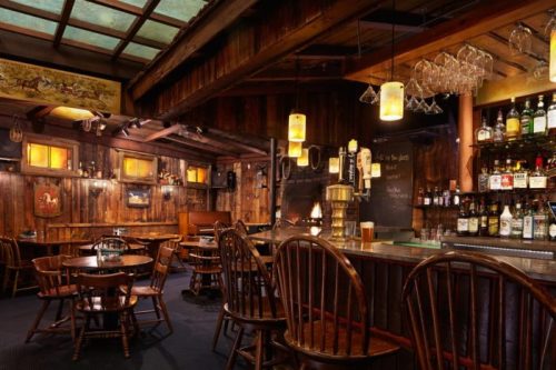 Cozy wooden bar interior with tables, a well-stocked bar, and warm lighting, creating a rustic atmosphere.