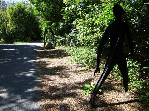 Two abstract sculptures resembling human figures stand along a tree-lined path, surrounded by greenery.