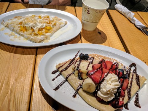 Two plates of crepes on a wooden table: one with fruit and whipped cream, the other with mango and almonds, plus a drink.