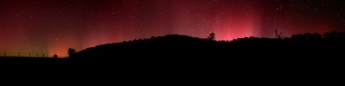 A panoramic view of a dark landscape illuminated by vibrant red and pink auroras against a starry night sky.
