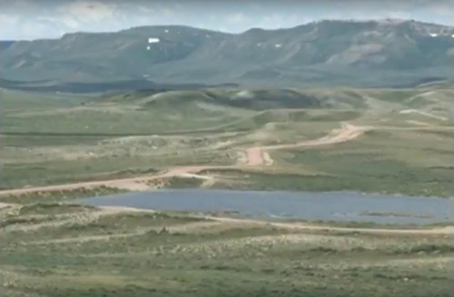 A scenic landscape featuring rolling hills, a dirt road, and a small pond surrounded by greenery under a cloudy sky.