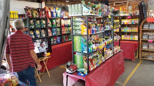 A market stall filled with various snacks and groceries, with a person browsing and a TV displaying a black-and-white film.