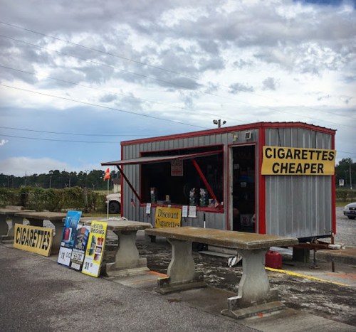 A small roadside stand with signs advertising cheaper cigarettes, surrounded by concrete benches and a cloudy sky.