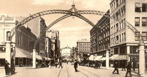 Historic street scene featuring ornate arches, shops, and pedestrians in a bustling urban setting.