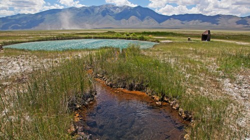 A serene landscape featuring a small pond and stream, surrounded by grass and mountains under a partly cloudy sky.