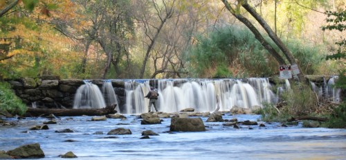 A serene waterfall cascades over rocks, with a person fishing in the foreground amidst autumn foliage.