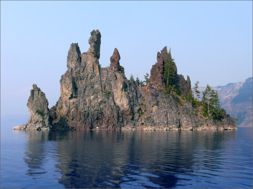 A rocky island with jagged formations and sparse trees, reflected in calm blue water under a clear sky.