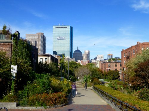 A scenic view of a city skyline with greenery and a pathway, featuring tall buildings under a clear blue sky.