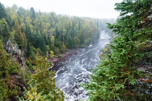A misty river flows through a forested landscape, surrounded by vibrant autumn foliage and rocky cliffs.