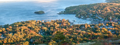 A scenic view of a coastal town surrounded by autumn foliage and a calm bay, with boats dotting the water.