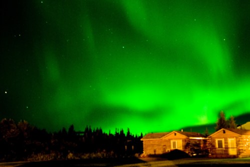 A vibrant green aurora borealis illuminates the night sky above rustic cabins and a forested landscape.