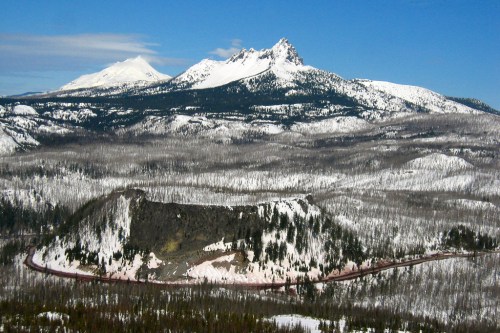 Snow-covered mountains rise above a forested landscape, with a winding road visible in the foreground.