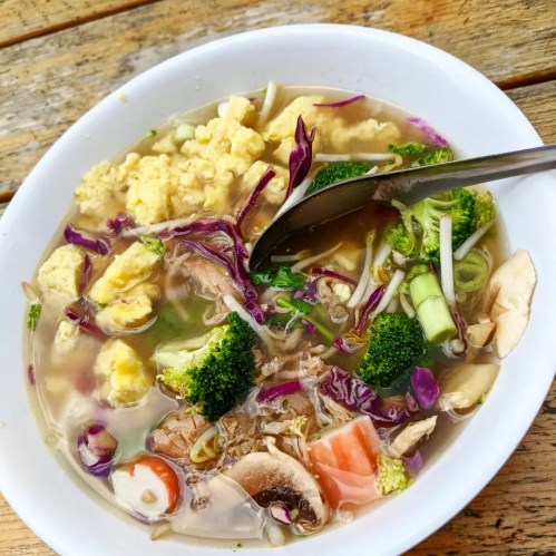 A bowl of soup with vegetables, chicken, and egg, served with a spoon on a wooden table.