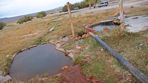 Two small hot springs surrounded by grass, with a pipe leading from one pool, and a vehicle parked in the background.