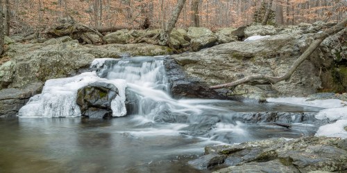 A serene waterfall cascades over rocks in a forest, surrounded by autumn foliage and a tranquil stream.