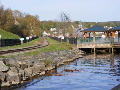 A peaceful waterfront scene with a wooden gazebo, rocky shoreline, and train tracks leading into the distance.