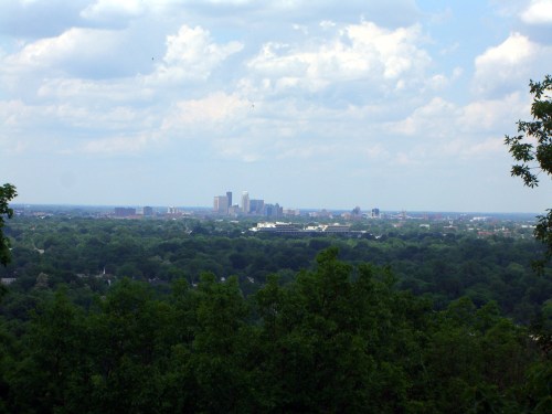 A panoramic view of a city skyline surrounded by lush green trees under a cloudy sky.