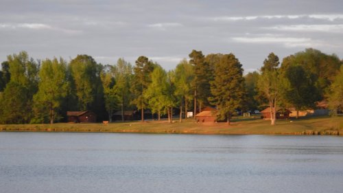 A serene lakeside view with green trees and rustic cabins along the shore under a cloudy sky.